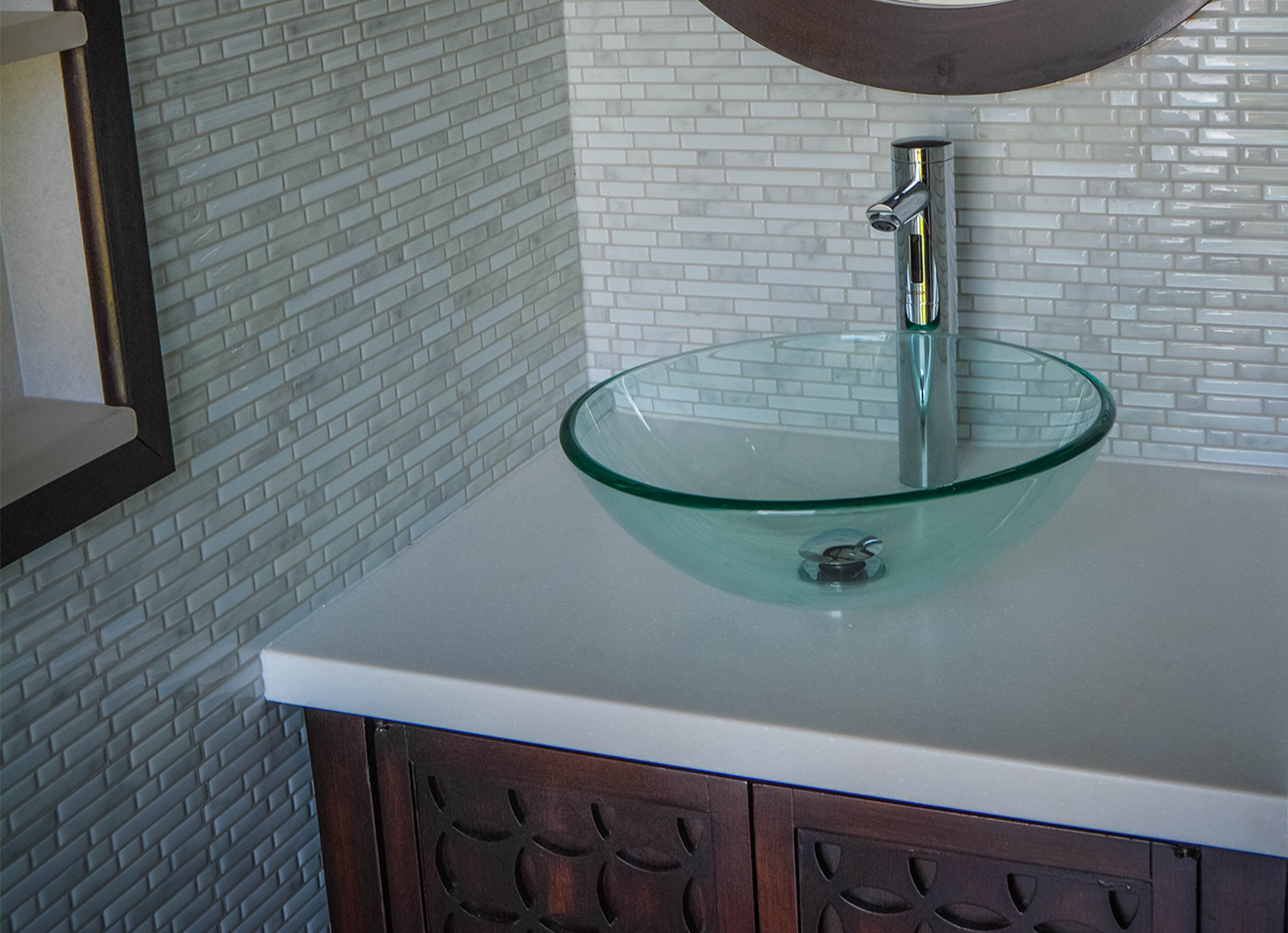 A close-up of a modern glass bowl sink resting on a white countertop atop a wooden cabinet; the sink features a sleek metallic silver faucet, with clean tile on the wall and a brown-framed mirror above, creating a stylish and contemporary bathroom design