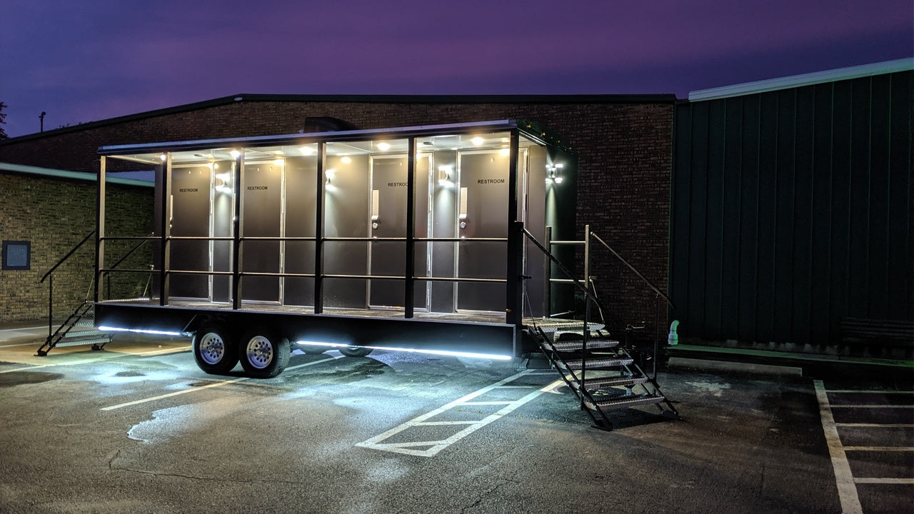 A grey restroom trailer with four stalls, photographed at night in a parking lot; each door is illuminated by individual overhead lights, with an additional strip of lighting underneath the trailer, casting a soft glow beneath it; the sky is a rich deep purple, adding atmosphere to the scene