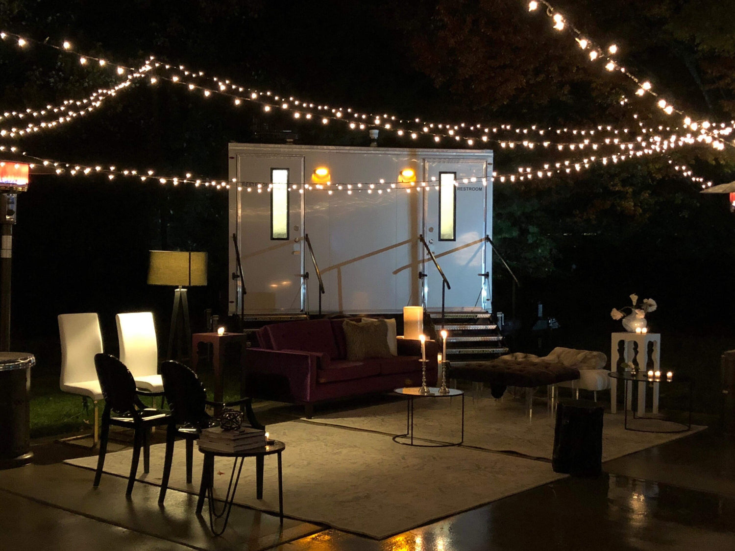 A cozy nighttime outdoor event setup featuring a clean white restroom trailer with two stalls, one on each side; the area is surrounded by chairs, glowing string lights, lit candles, lamp heaters, soft pillows, and layered rugs, creating a warm and inviting space ideal for a book reading or acoustic performance